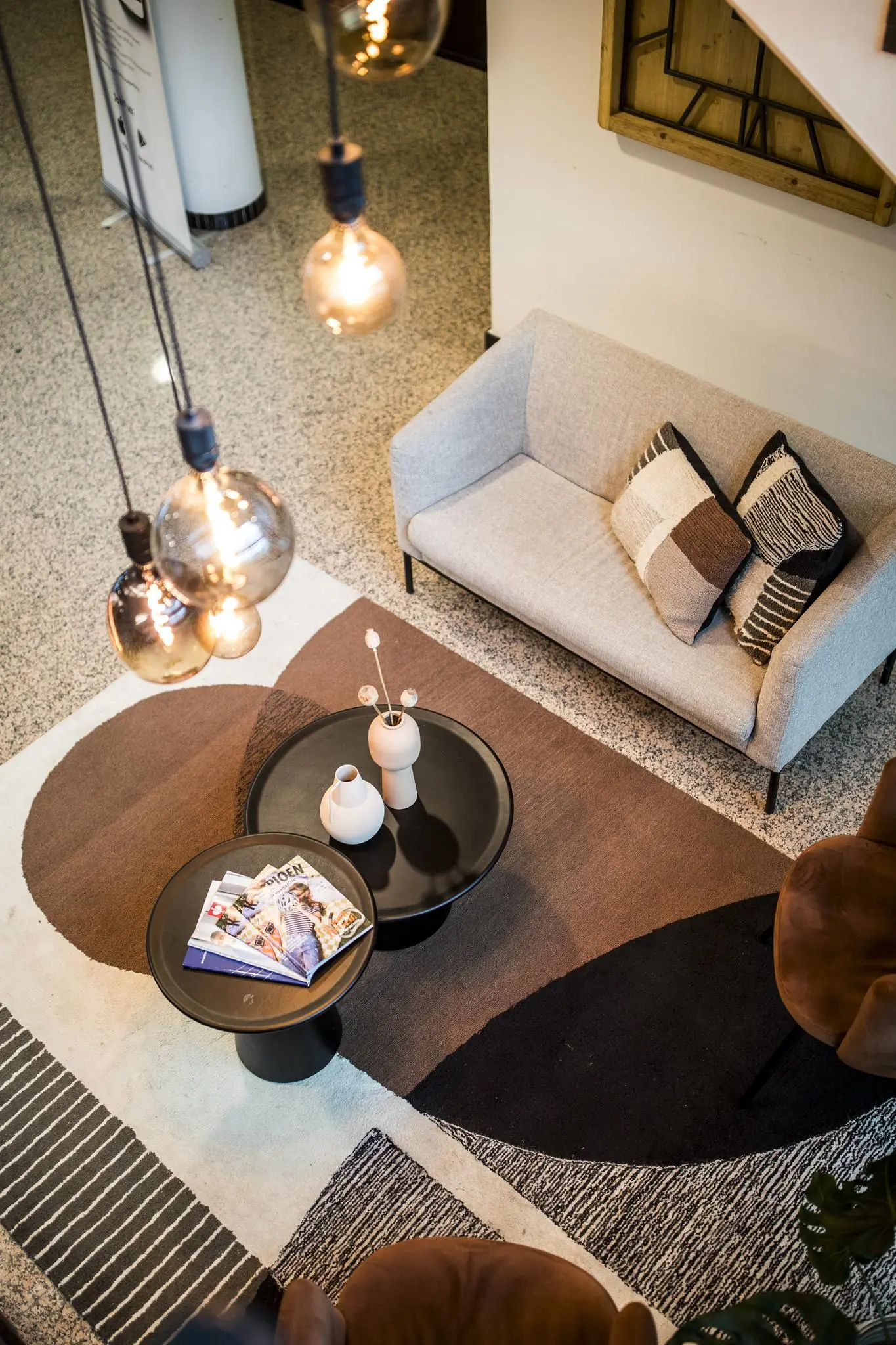 Moderne zithoek met een beige bank, designvloerkleed, twee zwarte ronde salontafels en decoratieve verlichting, gezien van bovenaf.